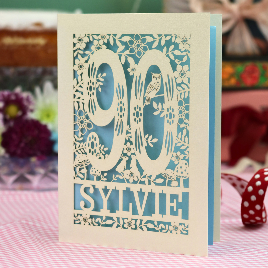 A handmade paper cut 90th birthday card with laser-cut woodland details, the number 90, flowers, and an owl over a light blue background, personalized with the name 'SYLVIE' at the bottom. The card is standing on a pink and white checked tablecloth with a soft-focus background featuring a 90th birthday gifts and a 90th birthday cake.