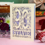 A laser-cut paper birthday card with floral and bird cutout details, featuring the number 90 and the name 'SYLVIE' on a pink and white checkered tablecloth with decorative flowers and ribbons in the background.