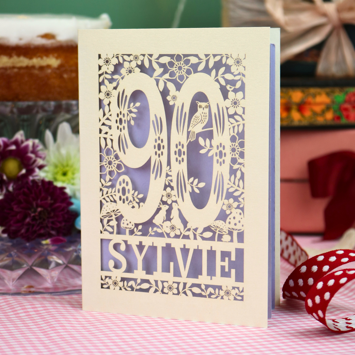 A laser-cut paper birthday card with floral and bird cutout details, featuring the number 90 and the name 'SYLVIE' on a pink and white checkered tablecloth with decorative flowers and ribbons in the background.