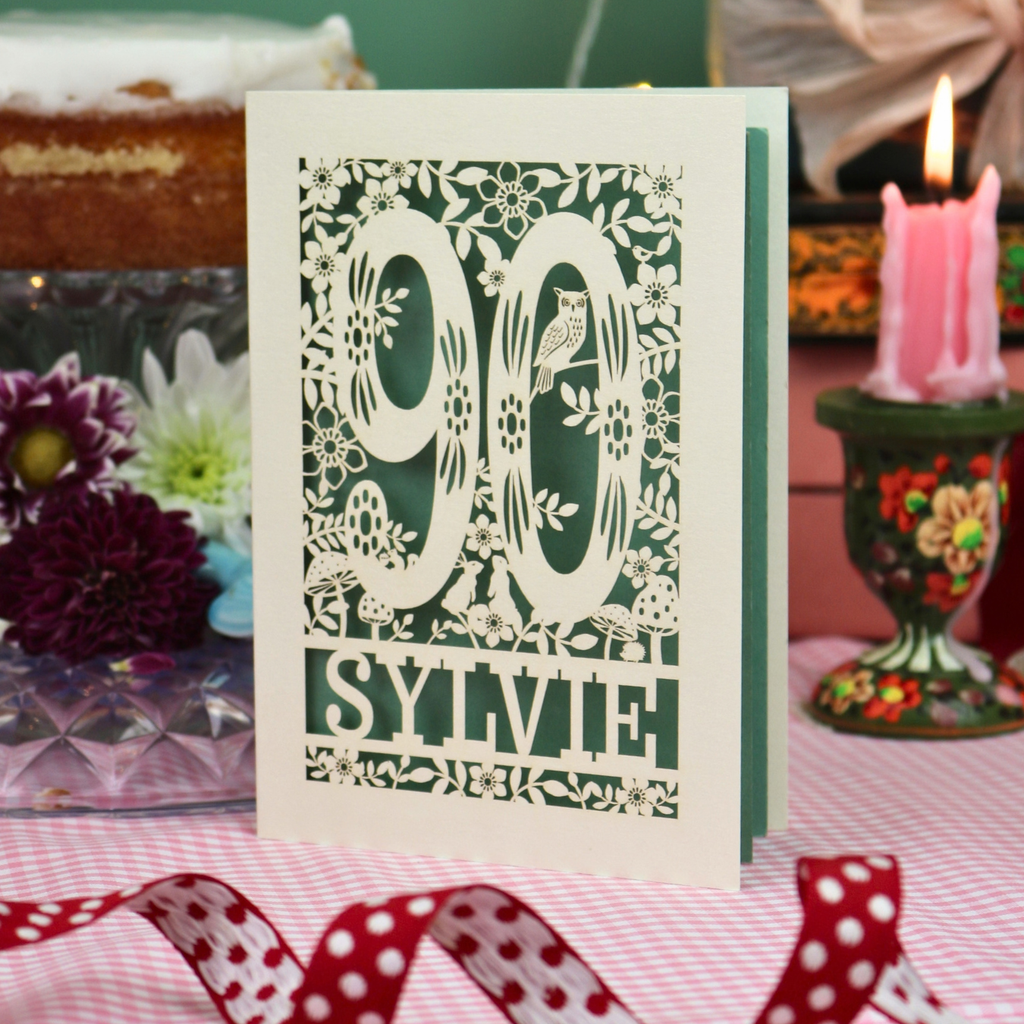 A handmade laser cut 90th birthday card with laser-cut woodland details, the number 90, flowers, and an owl over a sage background, personalized with the name 'SYLVIE' at the bottom. The card is standing on a pink and white checked tablecloth with a soft-focus background featuring a 90th birthday gifts and a 90th birthday cake.