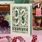 A handmade laser cut 90th birthday card with laser-cut woodland details, the number 90, flowers, and an owl over a sage background, personalized with the name 'SYLVIE' at the bottom. The card is standing on a pink and white checked tablecloth with a soft-focus background featuring a 90th birthday gifts and a 90th birthday cake.