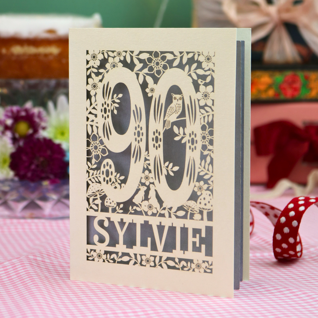 A personalized laser-cut greeting card for a 90th birthday, featuring the number 90 and the name 'SYLVIE' surrounded by floral and bird designs, standing on a pink and white checked tablecloth.