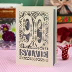 A personalized laser-cut greeting card for a 90th birthday, featuring the number 90 and the name 'SYLVIE' surrounded by floral and bird designs, standing on a pink and white checked tablecloth.