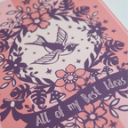 A pink A5 notebook with purple illustrations including a bird and flowers, and text "All of my Best Ideas"