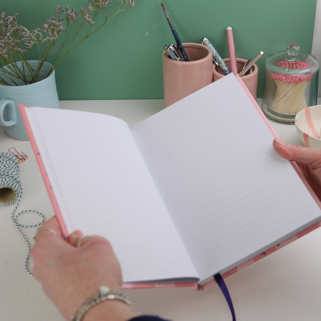 Open notebook held by a person with stationery items on a table in the background.