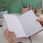 Open notebook held by a person with stationery items on a table in the background.