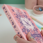 Person holding a pink notebook with purple illustrations on a light surface.