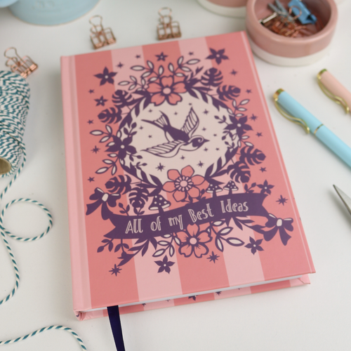 Pink A5 notebook with floral and bird design on a desk with stationery items