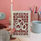 Valentine's Card with laser-cut design of 'Megan & Callum' held by a hand against a pink background.