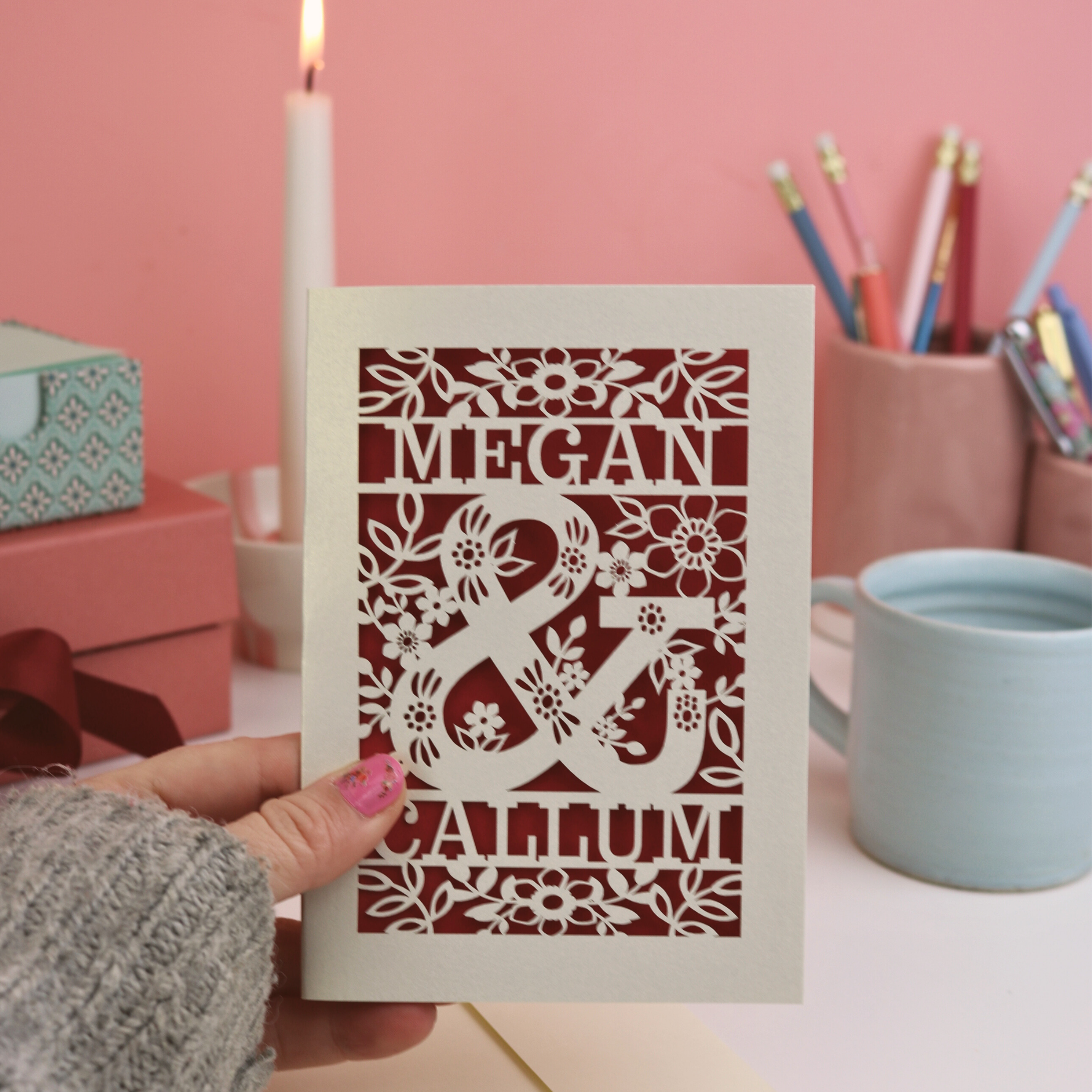 Valentine's Card with laser-cut design of 'Megan & Callum' held by a hand against a pink background.