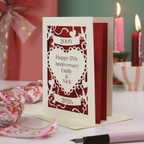 A laser cut personalised wedding anniversary card with 2009 cut into a banner, "Happy 17th Anniversary Emily & Nick" cut away from a heart shape, and 2026 in a banner underneath. Card is on a white surface with green background and candles, ribbon and a pen around it. 