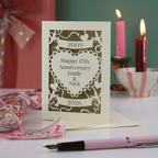 A laser cut personalised anniversary card with 2009 cut into a banner, "Happy 17th Anniversary Emily & Nick" cut away from a heart shape, and 2026 in a banner underneath. Card is on a white surface with green background and candles, ribbon and a pen around it. 