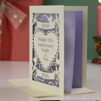 A laser cut personalised anniversary card with 2009 cut into a banner, "Happy 17th Anniversary Emily & Nick" cut away from a heart shape, and 2026 in a banner underneath. Card is on a white surface with green background and candles, ribbon and a pen around it. 