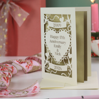 A paper cut personalised anniversary card with 2009 cut into a banner, "Happy 17th Anniversary Emily & Nick" cut away from a heart shape, and 2026 in a banner underneath. Card is on a white surface with green background and candles, ribbon and a pen around it. 