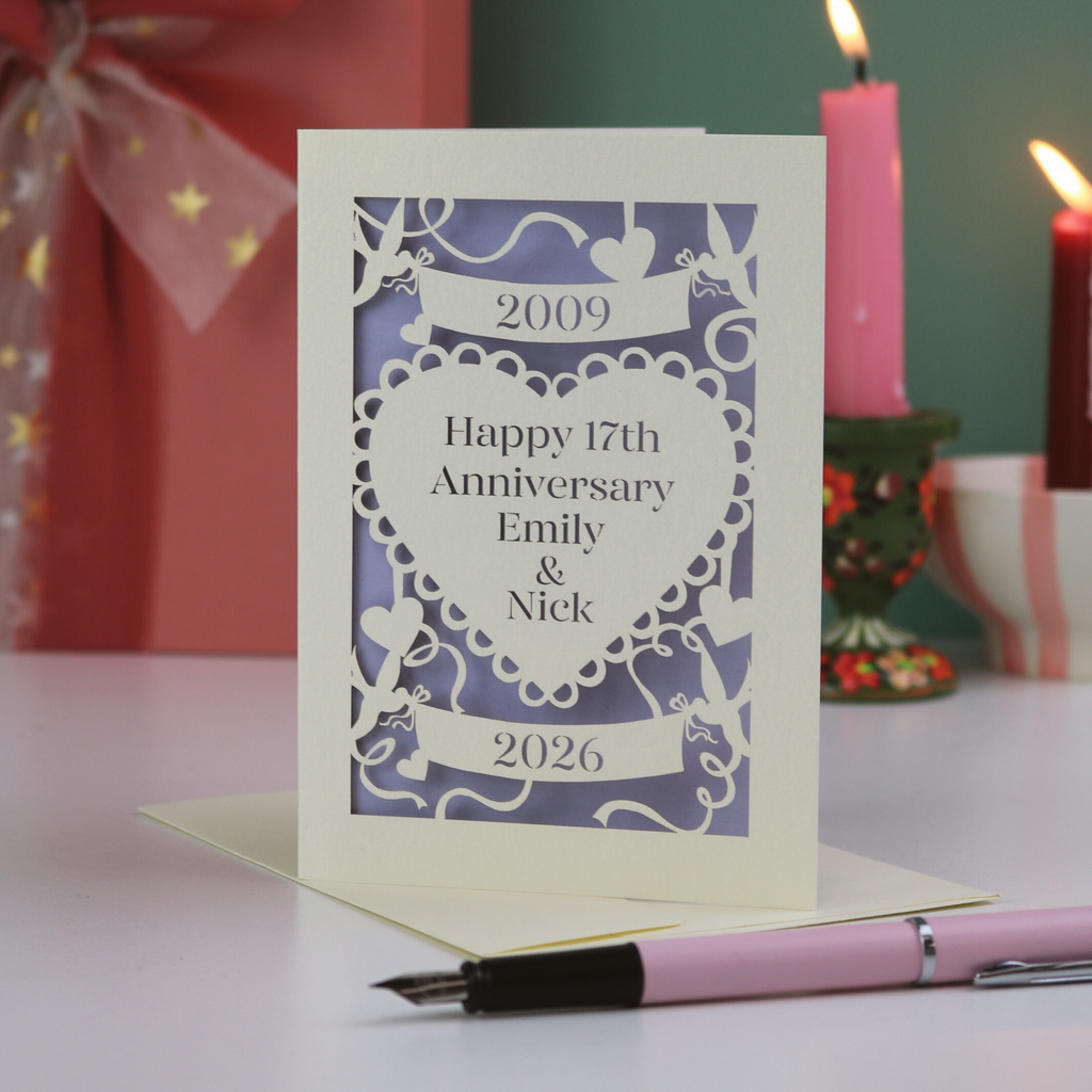 A laser cut personalised anniversary card with 2009 cut into a banner, "Happy 17th Anniversary Emily & Nick" cut away from a heart shape, and 2026 in a banner underneath. Card is on a white surface with green background and candles, ribbon and a pen around it. 
