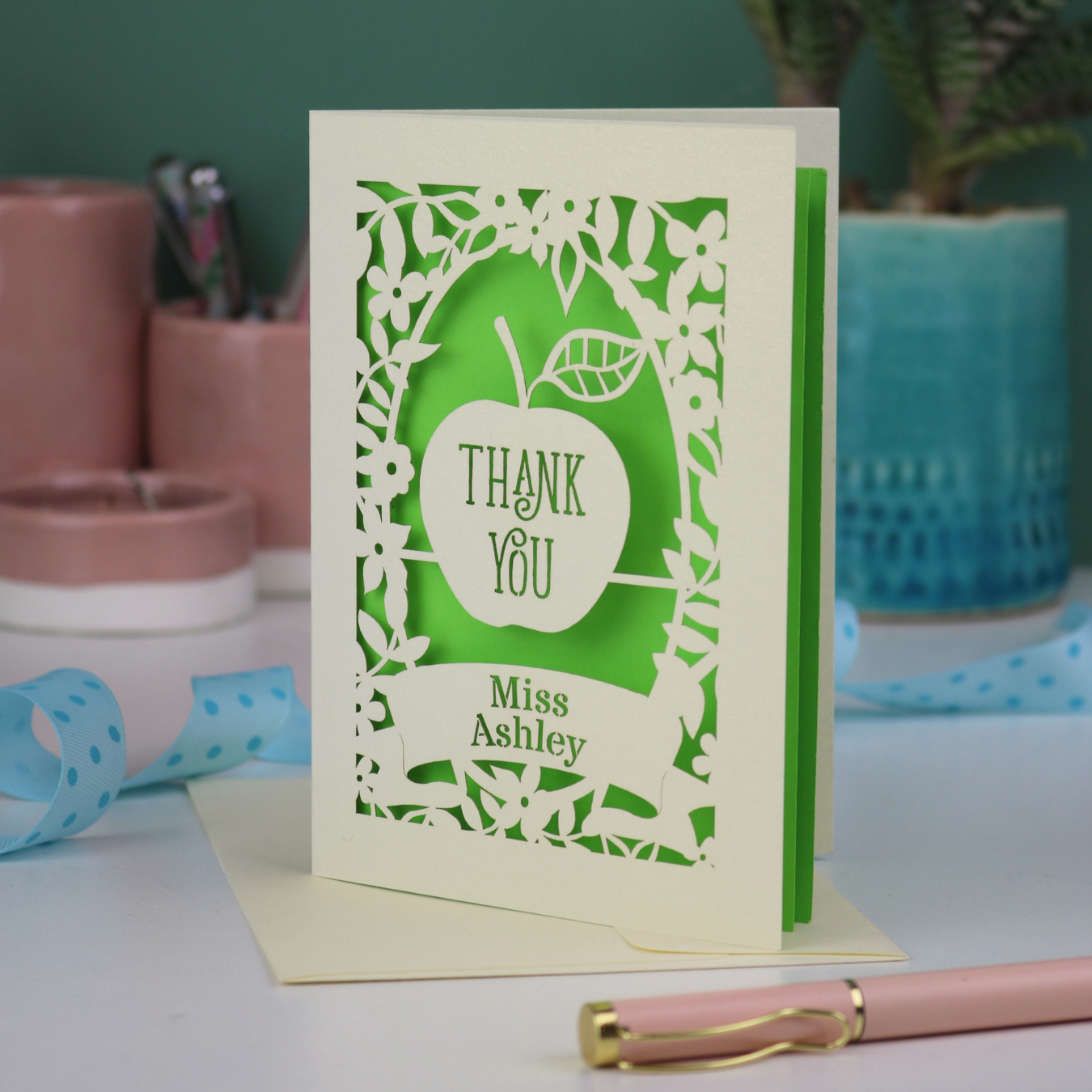 Personalised Thank you teacher card with apple design, decorative border and text, 'Thank You Miss Ashley',  on a desk with stationery items.