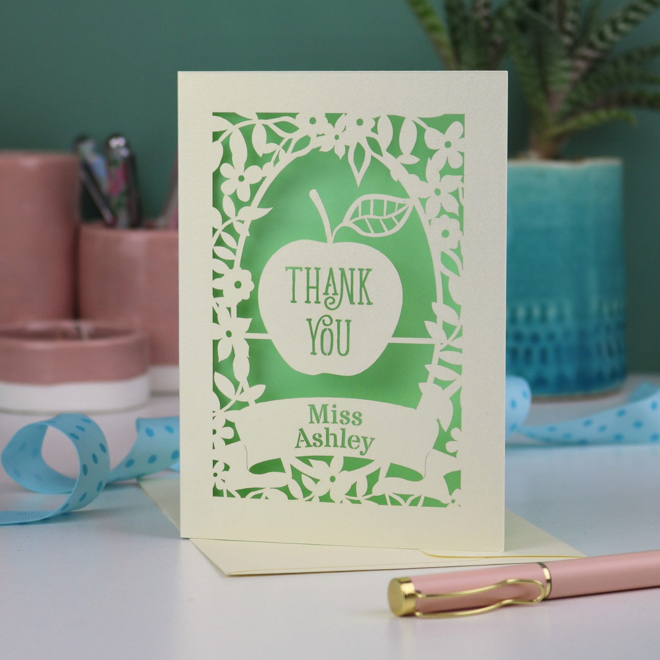 Paper cut Thank you teacher card with apple design, decorative border and text, 'Thank You Miss Ashley',  on a desk with stationery items.