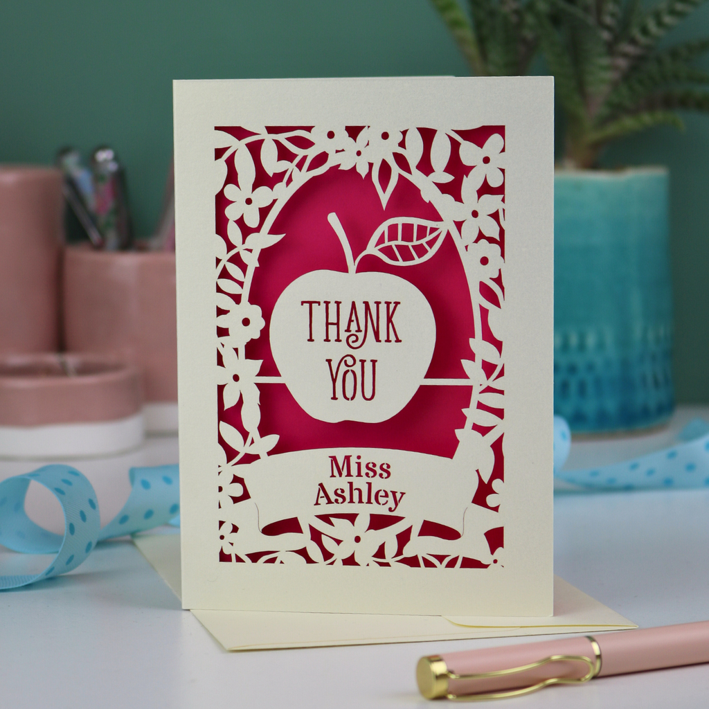 Thank you teacher card with apple design, decorative border and text, 'Thank You Miss Ashley',  on a desk with stationery items.
