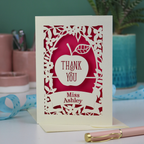 Thank you teacher card with apple design, decorative border and text, 'Thank You Miss Ashley',  on a desk with stationery items.