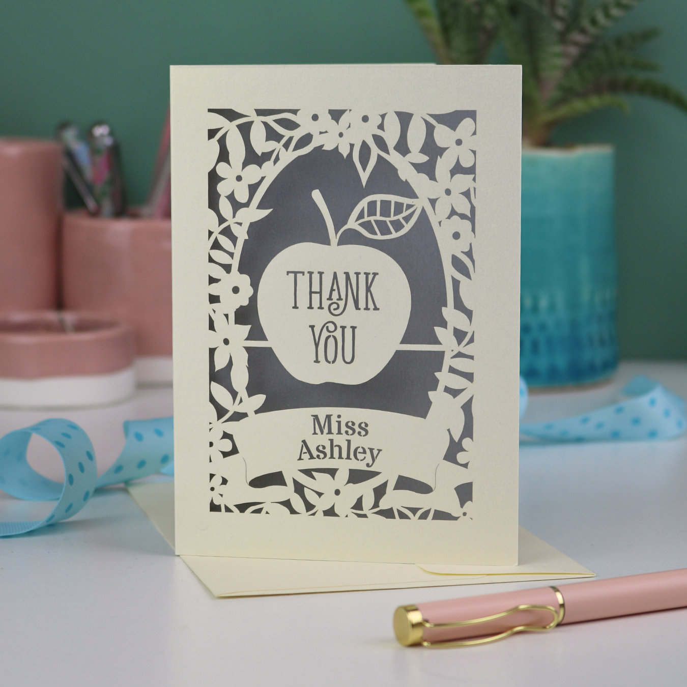 Personalized greeting card with 'Thank You Miss Ashley' text on a desk with stationery items.