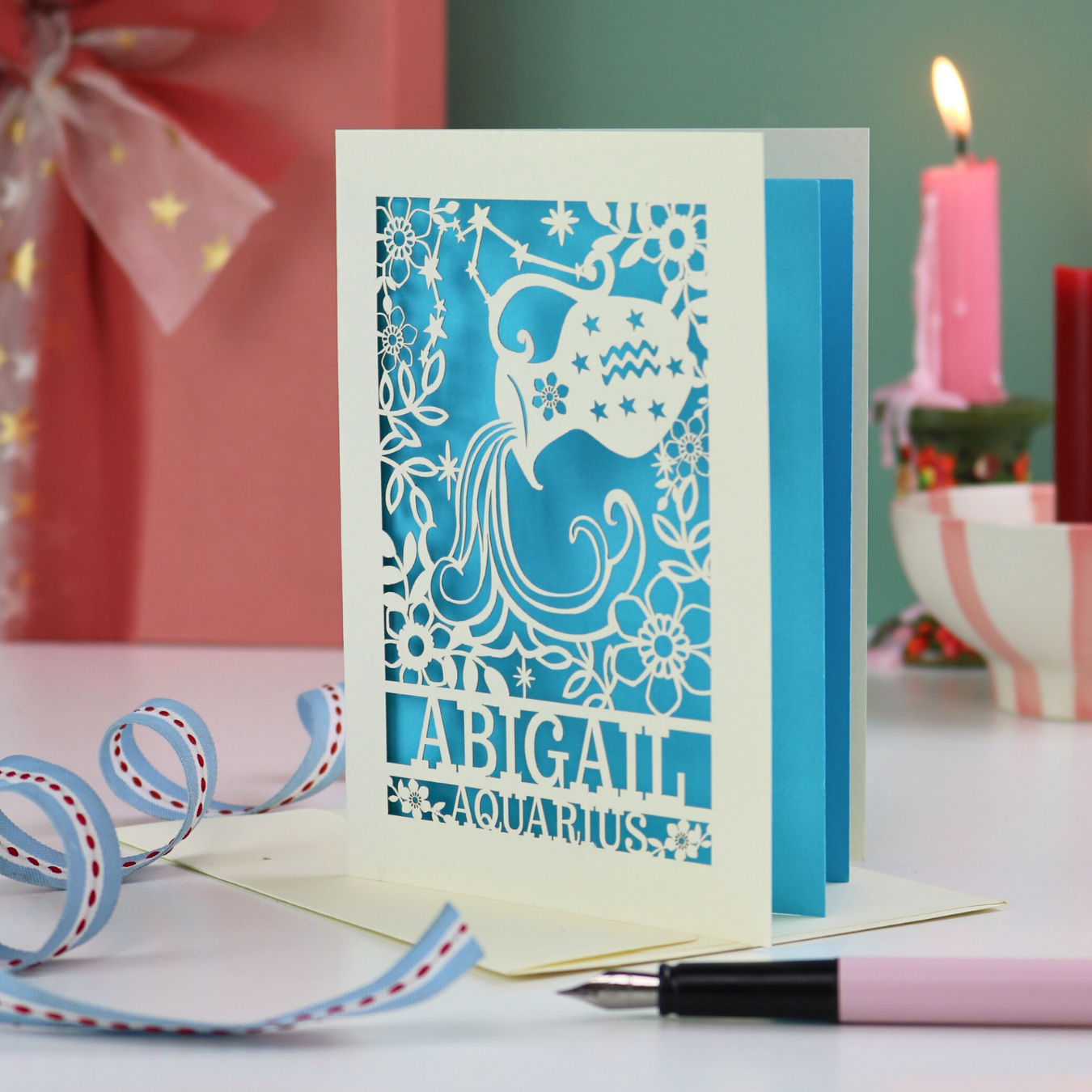A laser cut Aquarius birthday card personalised with the name Abigail, on a desk with candles and stationery items