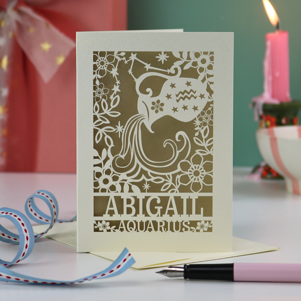 A laser cut Aquarius birthday card personalised with the name Abigail, on a desk with candles and stationery items