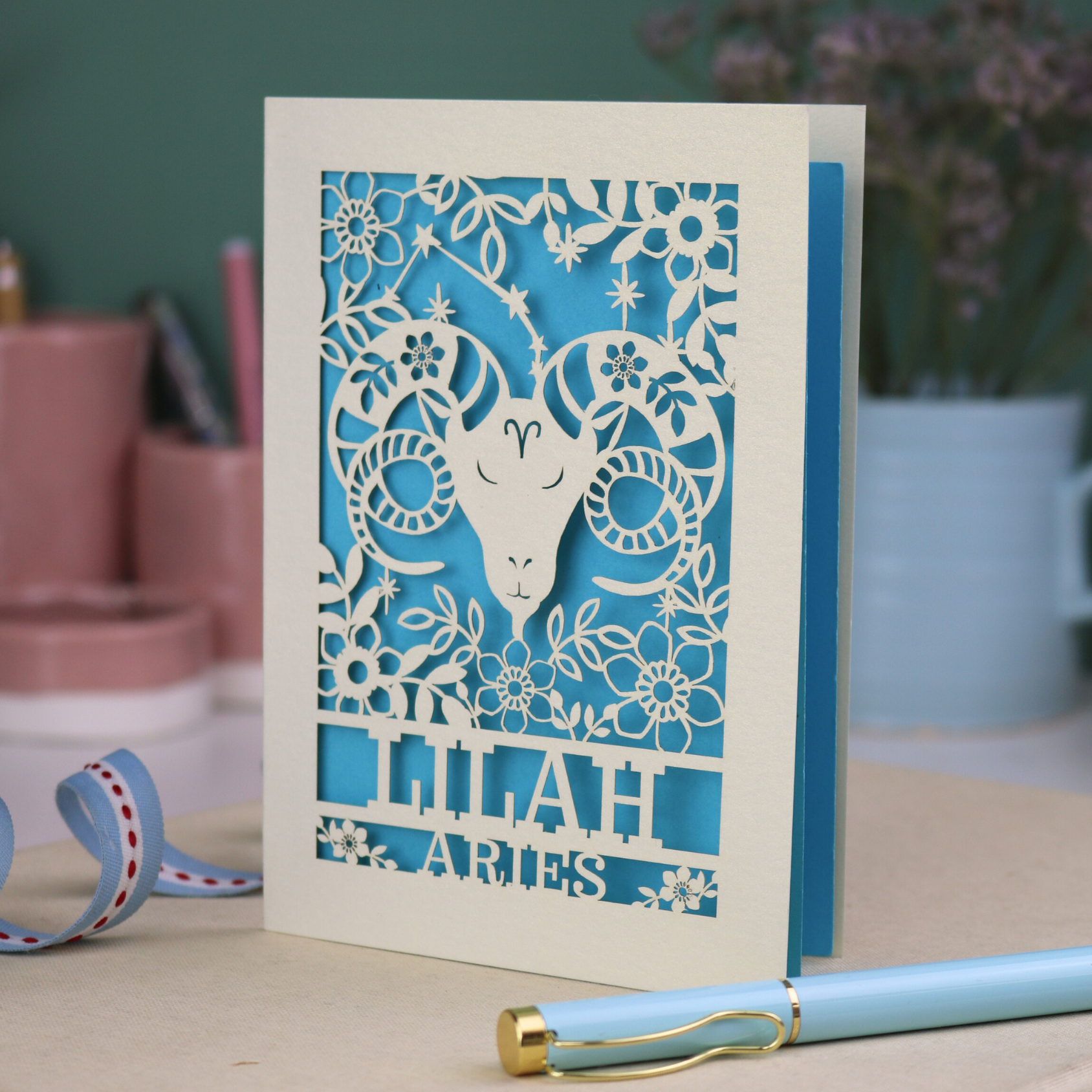 Aries birthday Card with laser-cut design of an Aries symbol and floral elements, with a pen and ribbons on a table.