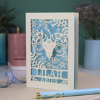 Aries birthday Card with laser-cut design of an Aries symbol and floral elements, with a pen and ribbons on a table.