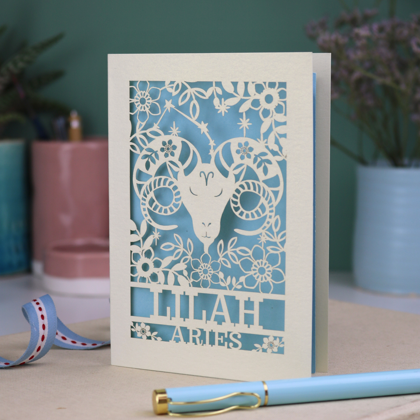 Aries birthday Card with laser-cut design of an Aries symbol and floral elements, with a pen and ribbons on a table.