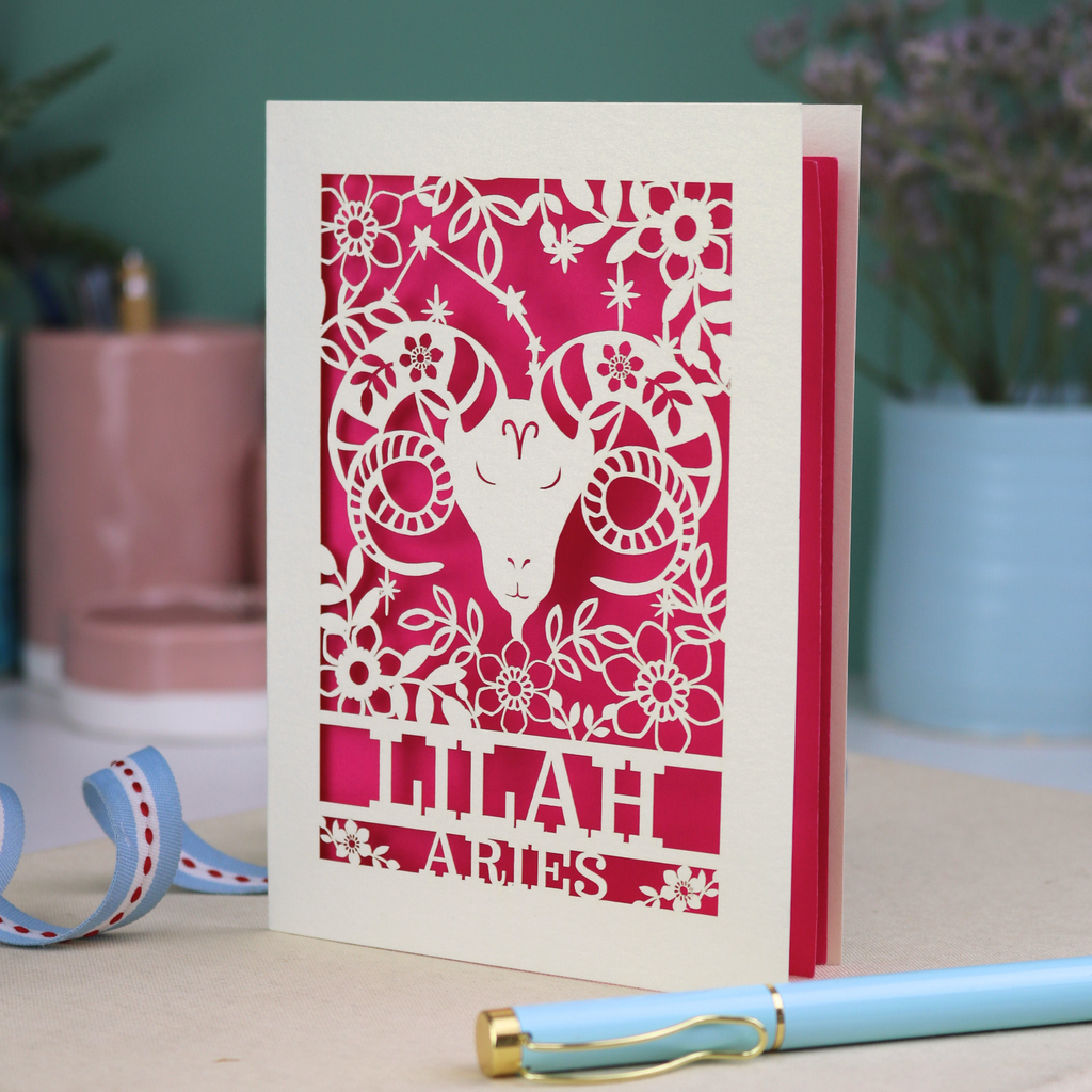 Birthday Card with zodiac design featuring an Aries symbol on a pink background, placed on a surface with decorative elements.