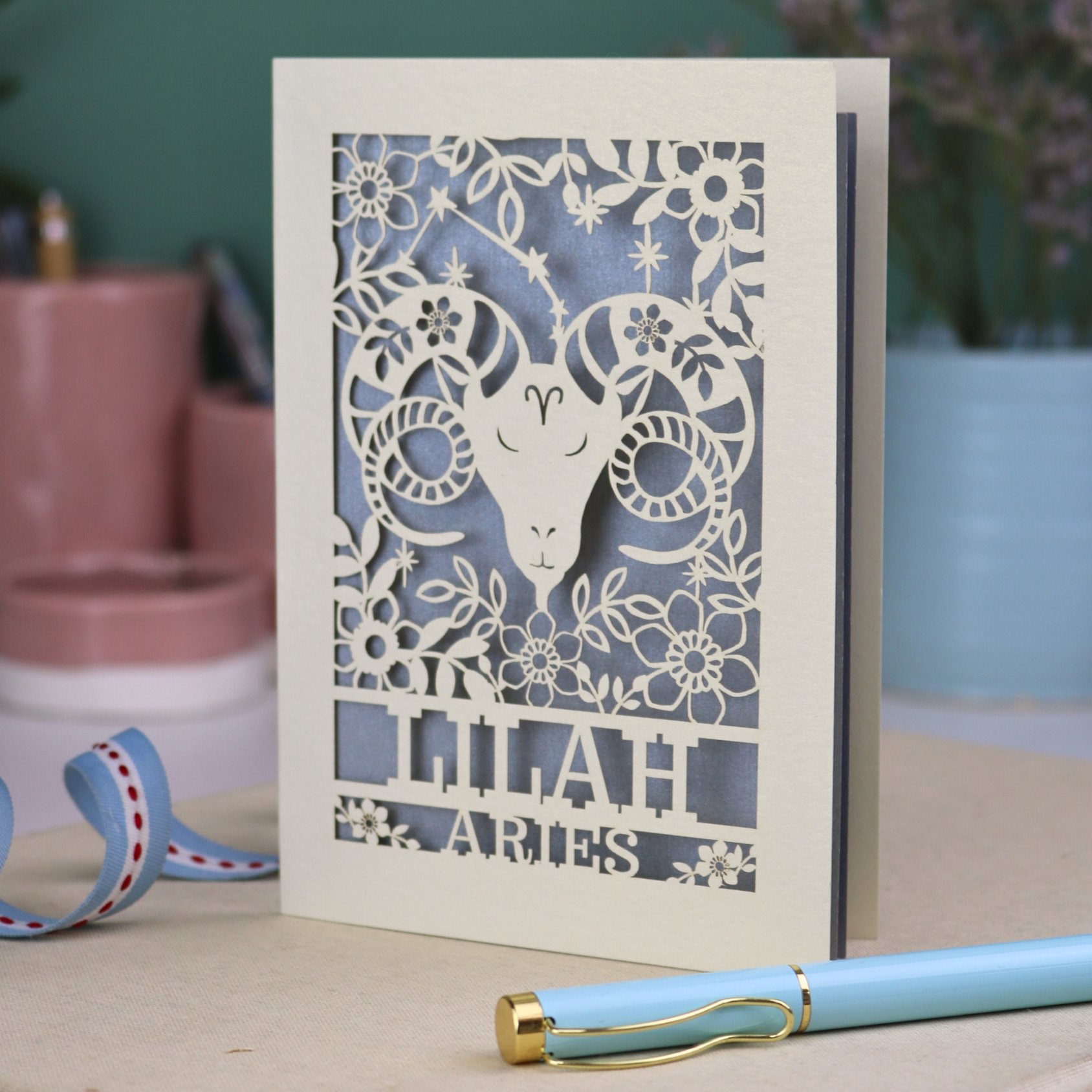 Aries birthday Card with laser-cut design of an Aries symbol and floral elements, with a pen and ribbons on a table.