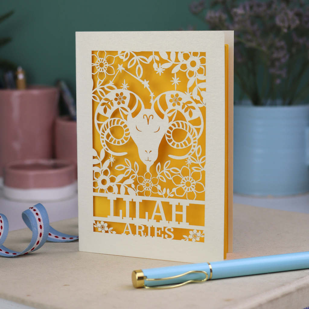 Aries birthday Card with laser-cut design of an Aries symbol and floral elements, with a pen and ribbons on a table.