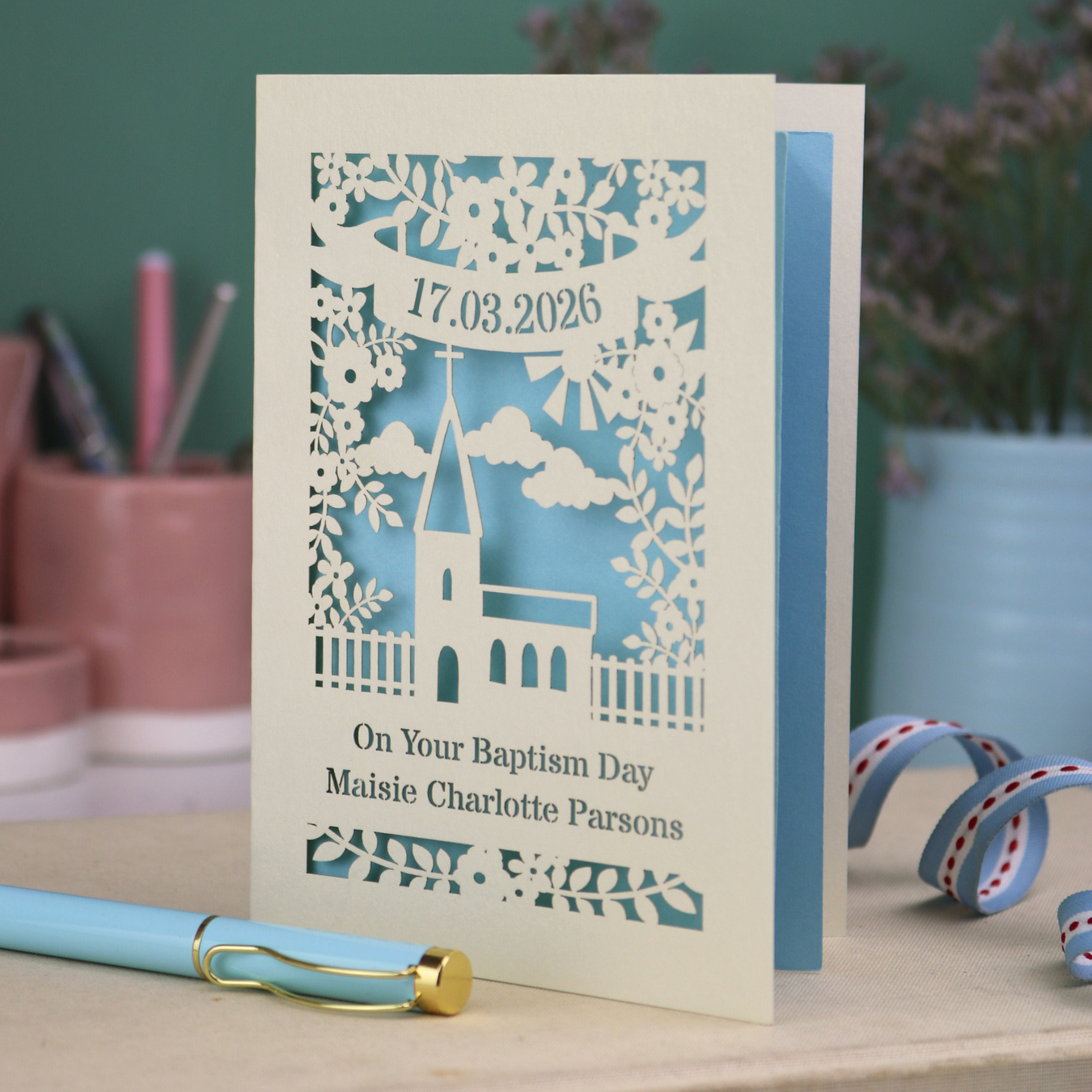 Personalized baptism card with floral church design and decorative elements on a table with stationery items.