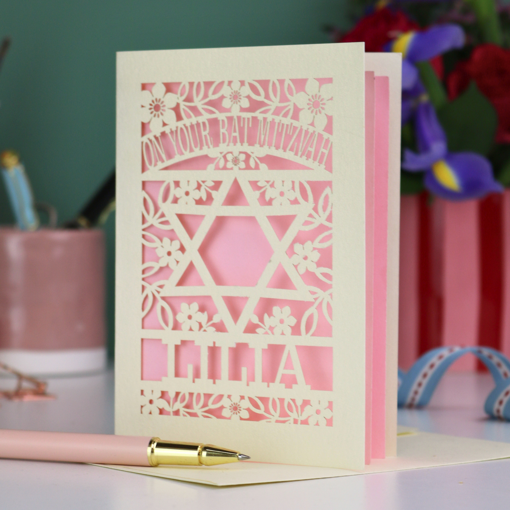 Cut out Bat Mitzvah card with decorative design and text on a table with flowers and stationery.