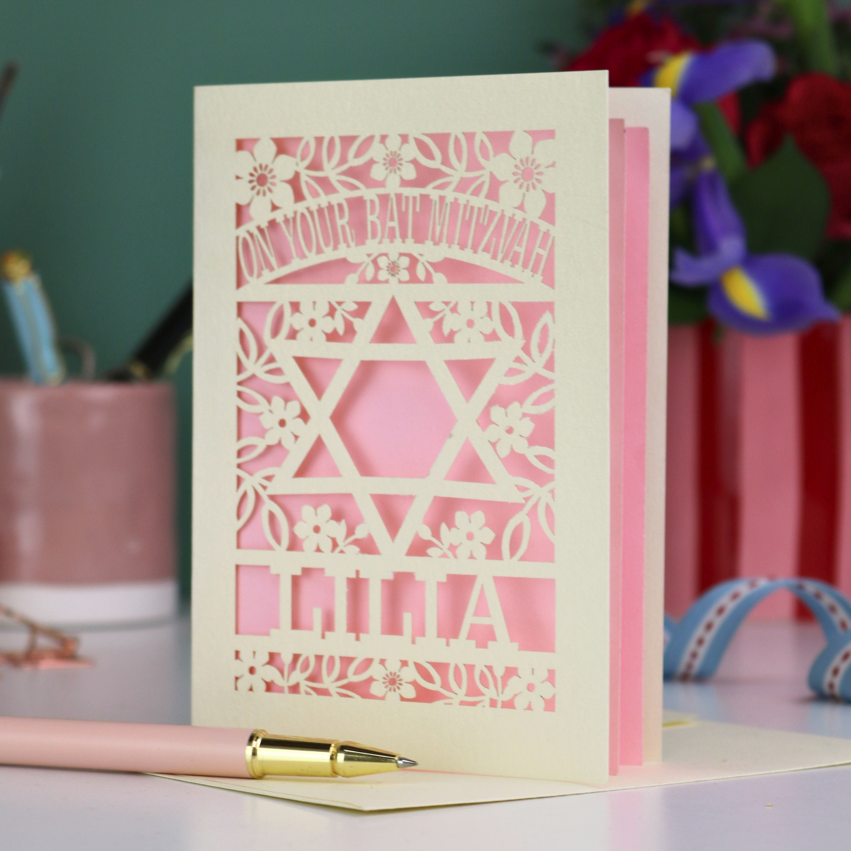 Cut out Bat Mitzvah card with decorative design and text on a table with flowers and stationery.