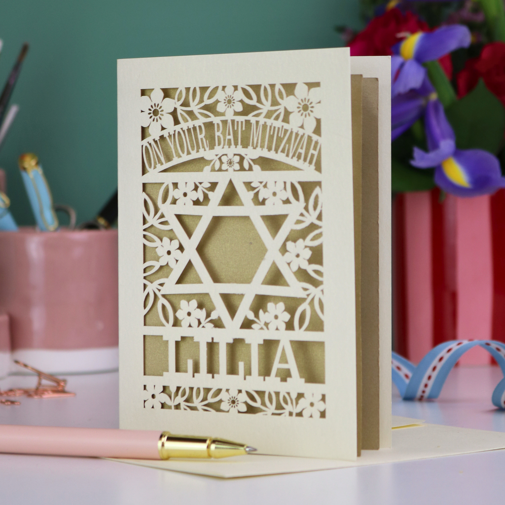 Personalised Laser-cut Bat Mitzvah card with decorative design and text on a table with flowers and stationery.