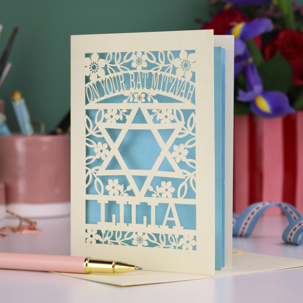 Laser-cut Bat Mitzvah card with decorative design and text on a table with flowers and stationery.