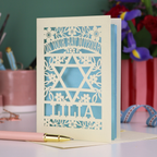 Laser-cut Bat Mitzvah card with decorative design and text on a table with flowers and stationery.