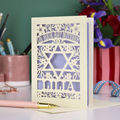 Laser-cut Bat Mitzvah card with decorative design and text on a table with flowers and stationery.