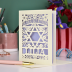 Laser-cut Bat Mitzvah card with decorative design and text on a table with flowers and stationery.