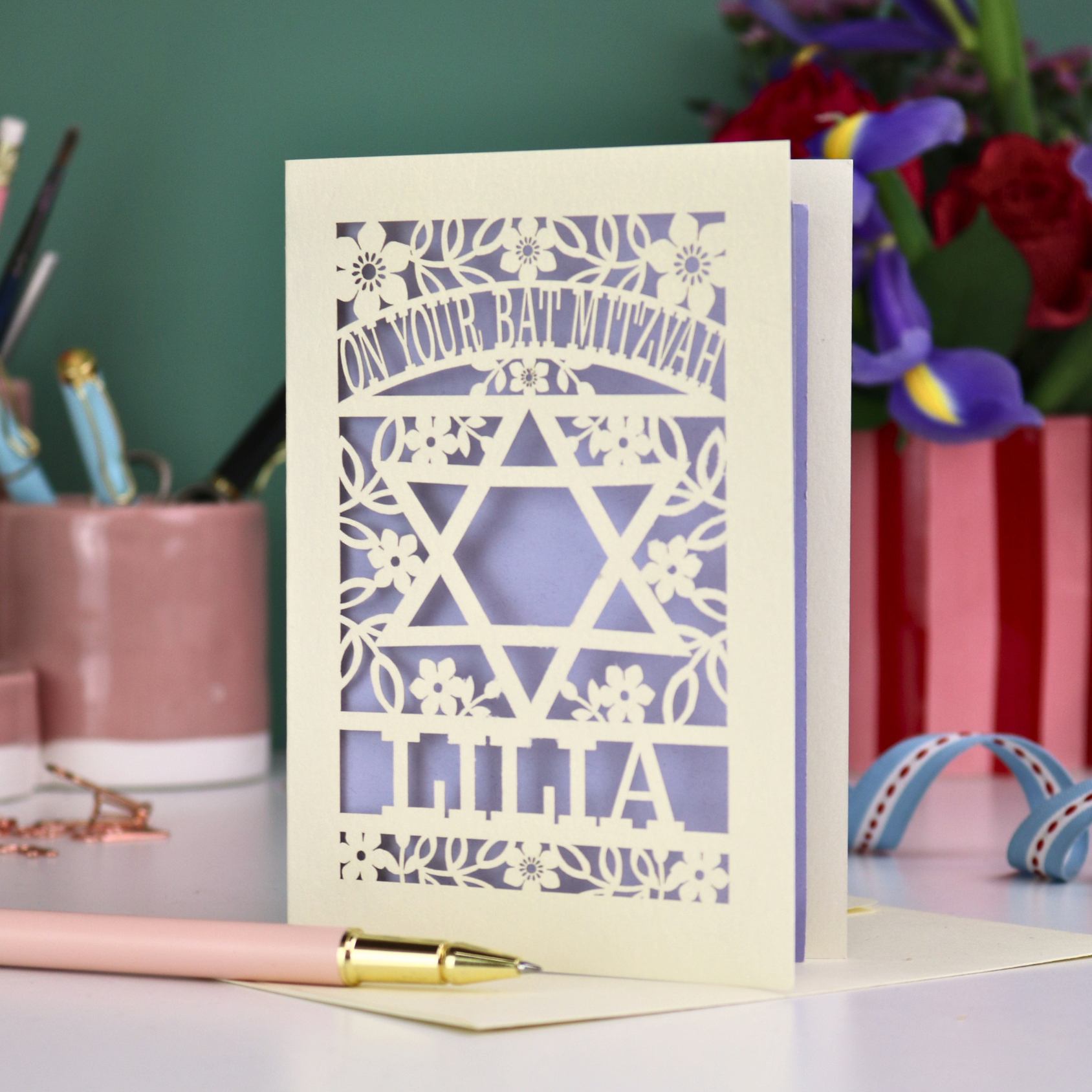 Laser-cut Bat Mitzvah card with decorative design and text on a table with flowers and stationery.