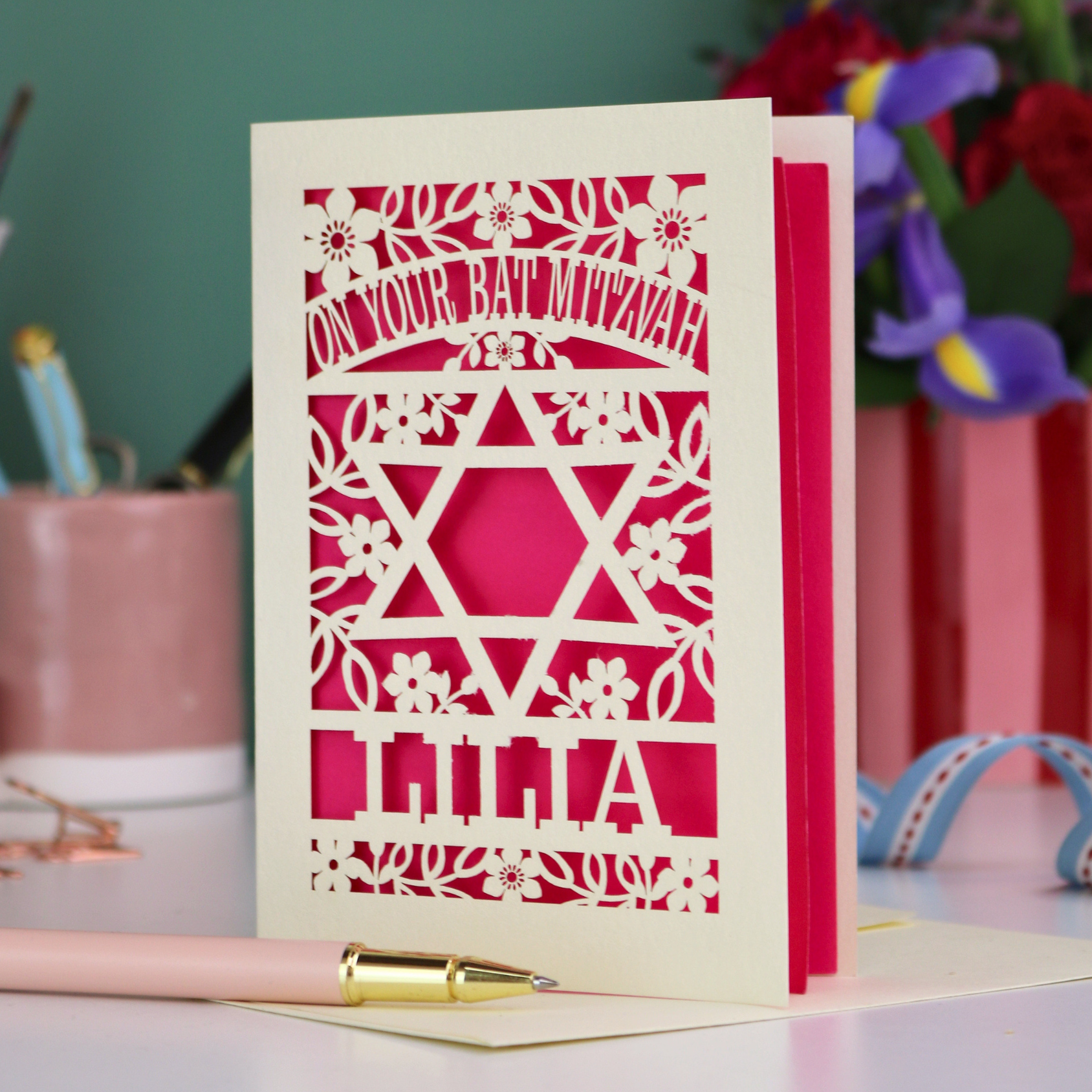 Laser-cut Bat Mitzvah card with Star of David and floral design on a desk with stationery items.