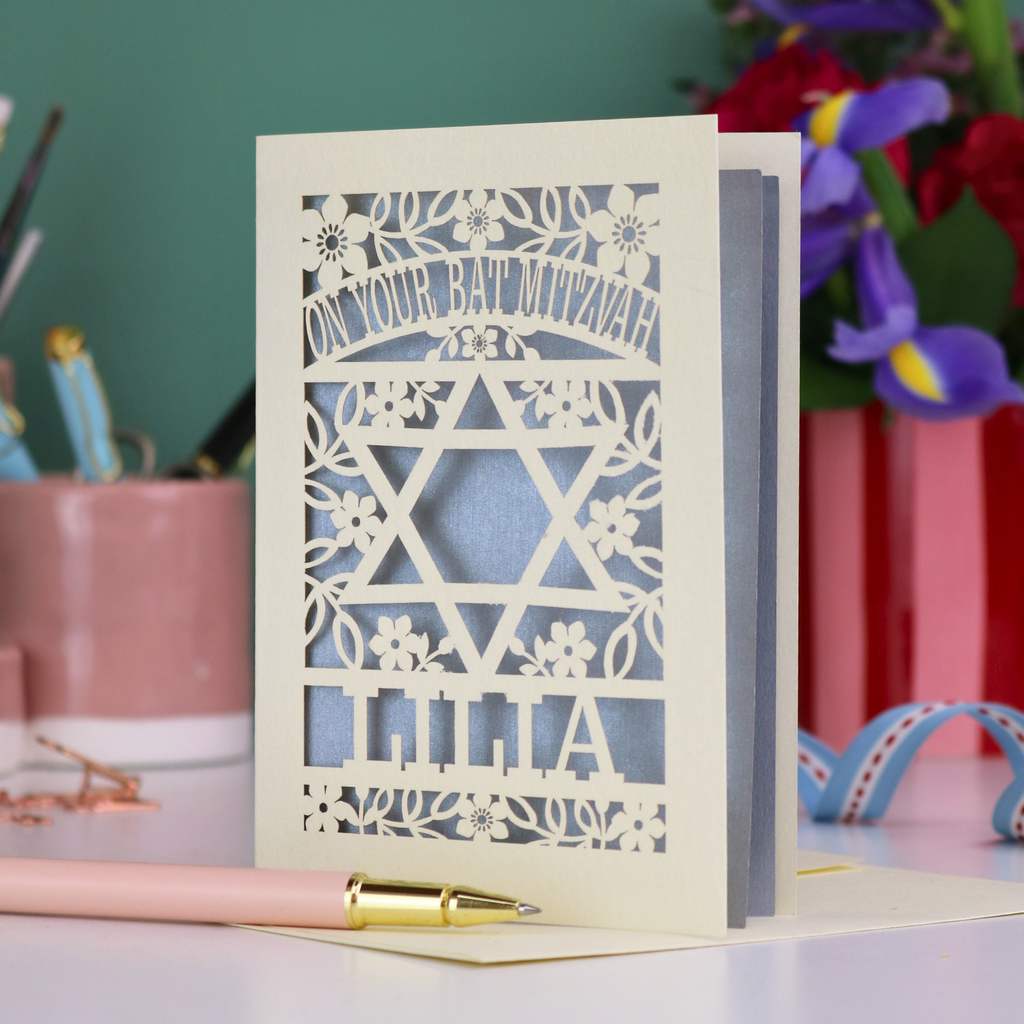 Special Laser cut Bat Mitzvah card with decorative design and text on a table with flowers and stationery.