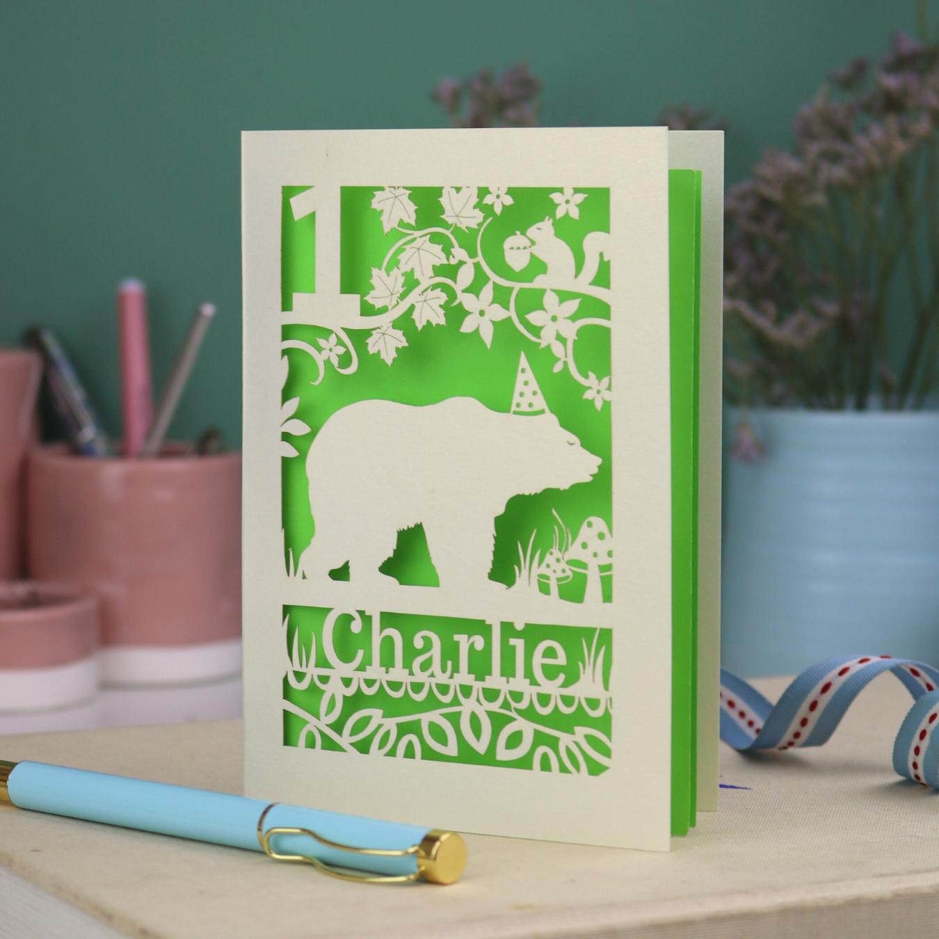 Laser cut 1st birthday card with bear design and 'Charlie' text on a table with stationery items.