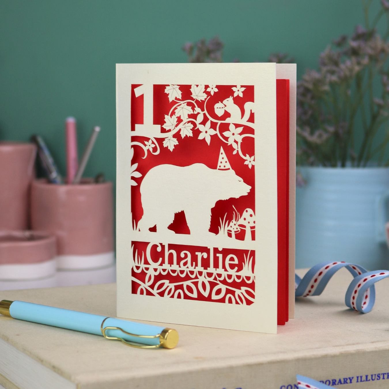 Paper cut first birthday card with bear design and 'Charlie' text on a table with stationery items.