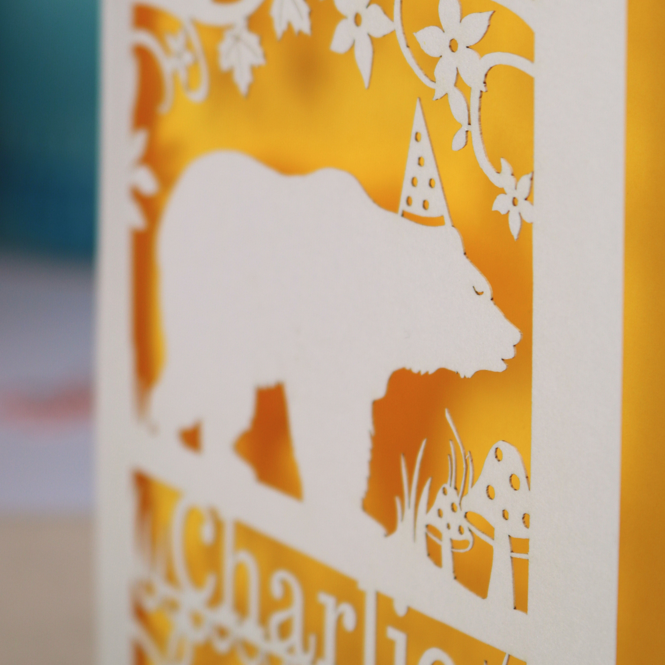 Paper cut 1st birthday card with bear design and 'Charlie' text on a table with stationery items.