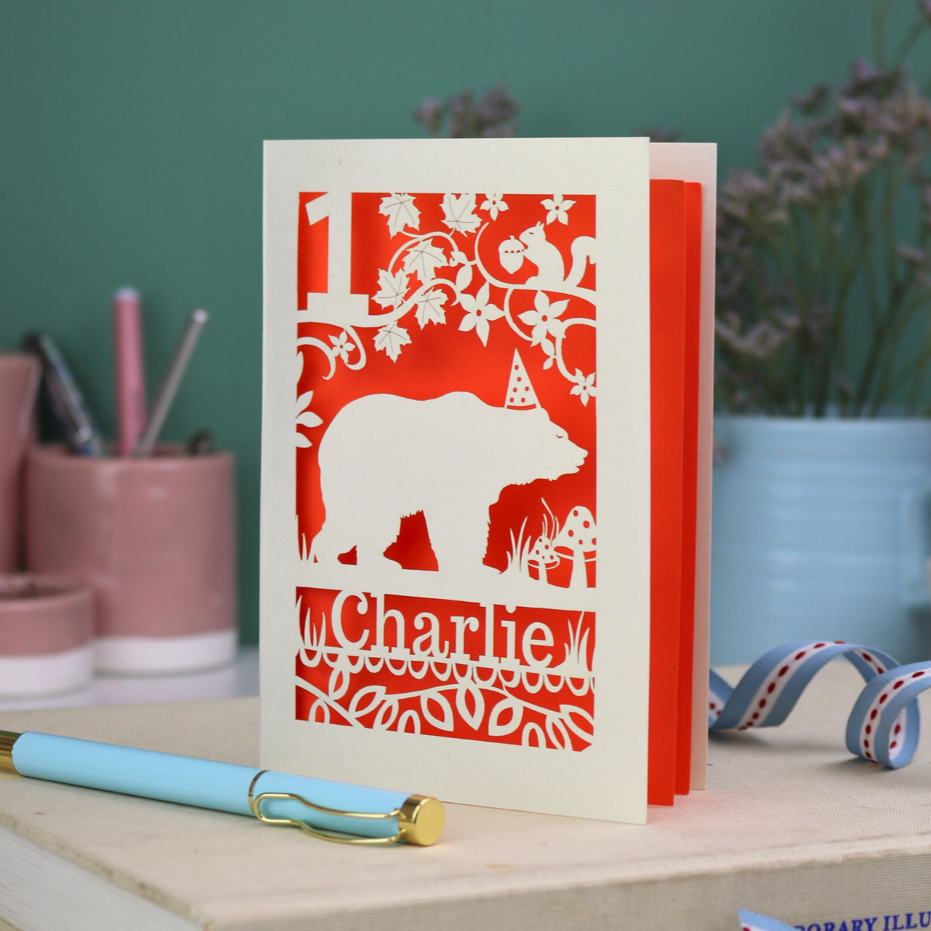 Personalised first birthday card with bear design and 'Charlie' text on a table with stationery items.