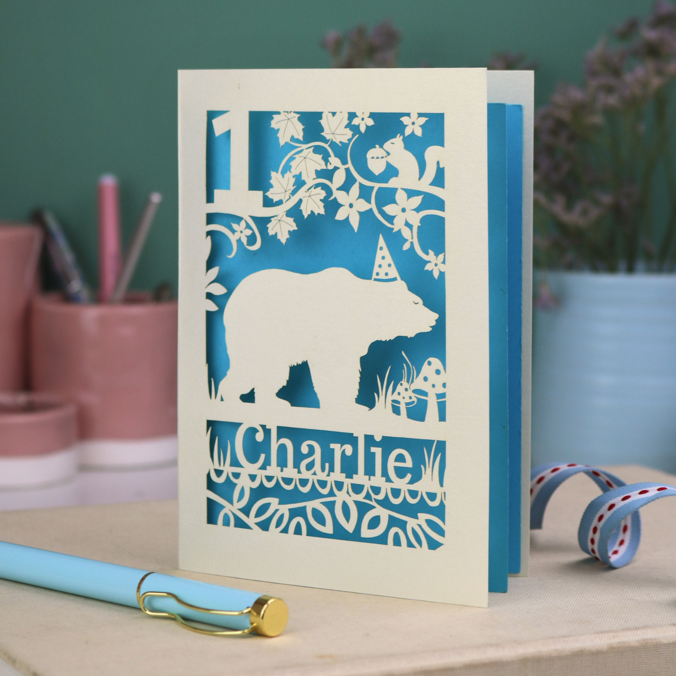 Laser cut first birthday card with bear design and 'Charlie' text on a table with stationery items.