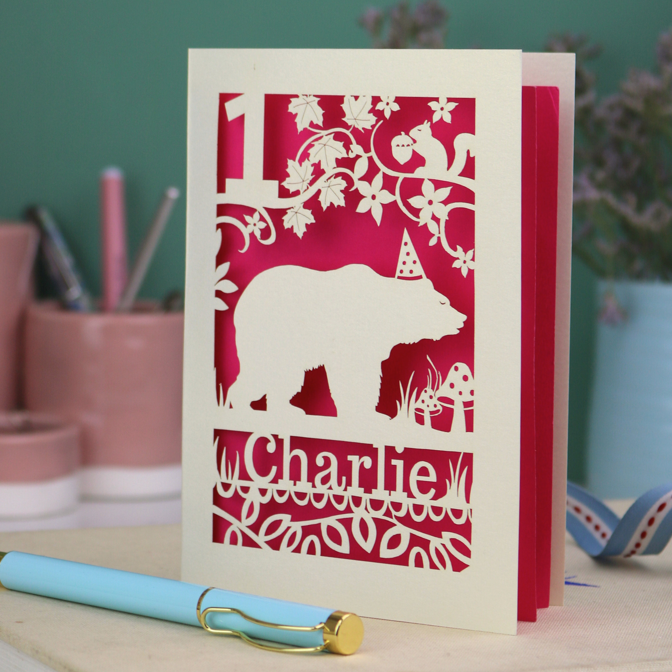 Personalised 1st birthday card with bear design and 'Charlie' text on a table with stationery items.