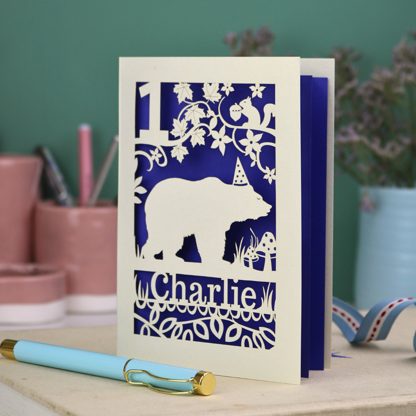 Personalised cut out first birthday card with bear design and 'Charlie' text on a table with stationery items.